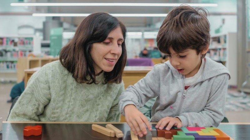 Une mère regarde son enfant assise à côté d'elle qui joue avec des blocs dans une bibliothèque.