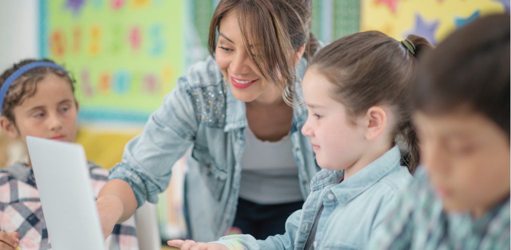 Une enseignante entourée de trois enfants montre à une petite fille quelque chose sur l'écran d'un ordinateur.