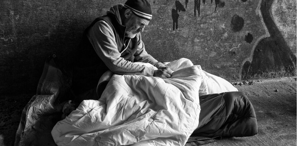 Photo en noir et blanc d'un homme en situation d'intinérance dans la rue avec son sac de couchage. 