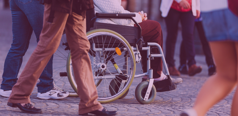Une personne en fauteuil roulant est entourée de personnes marchant dans une rue pavée. Seules leurs jambes et la partie inférieure de leur corps sont visibles.