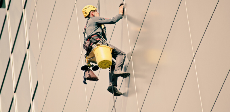 Un ouvrier portant un casque et un harnais est suspendu par des cordes alors qu'il nettoie la façade en verre d'un grand bâtiment. Il porte un grand seau jaune et des outils de nettoyage.