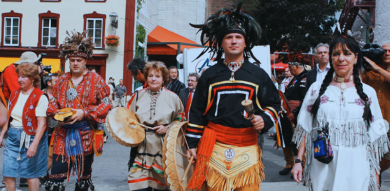 Un groupe de personnes, dont certaines portent des costumes traditionnels autochtones avec des plumes, des tambours et des perles, marchent ensemble dans un défilé, aux côtés d'autres personnes portant des vêtements modernes. Des bâtiments et des spectateurs sont visibles à l'arrière-plan.