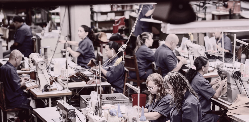 Un groupe de personnes en uniforme bleu travaille sur des machines à coudre dans une usine ou un atelier en pleine activité, entouré d'outils, de matériaux et d'étagères remplies de fournitures. 